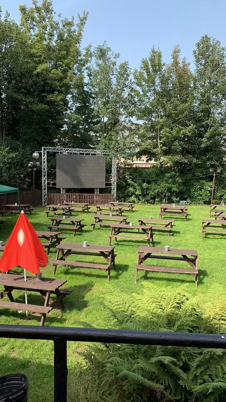 The garden by day: rows of wooden picnic benches on a mown lawn, with a large screen mounted on a black truss frame against the trees.
