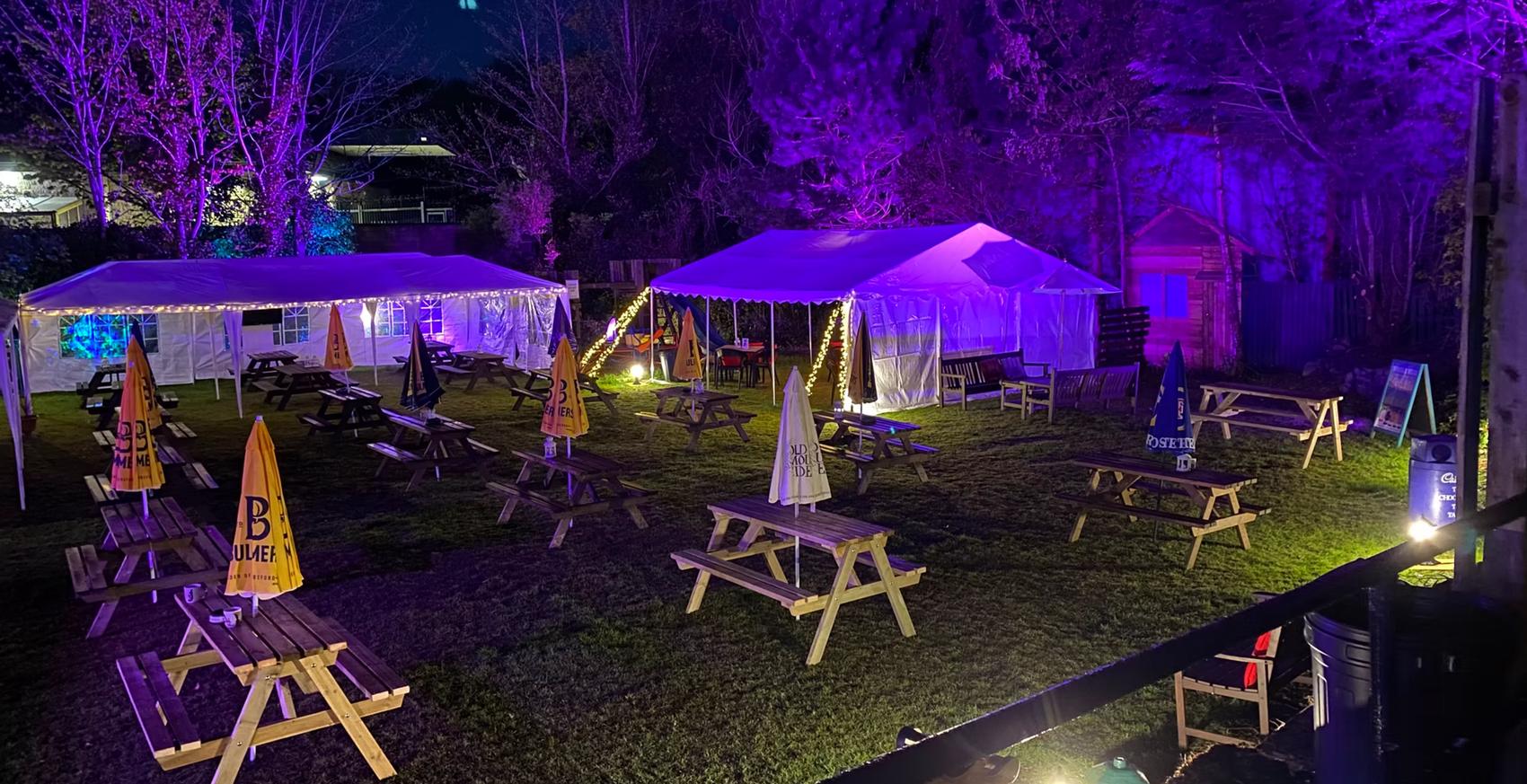 The Plough's garden at night: two marquees strung with festoon lights over rows of picnic benches on the lawn.